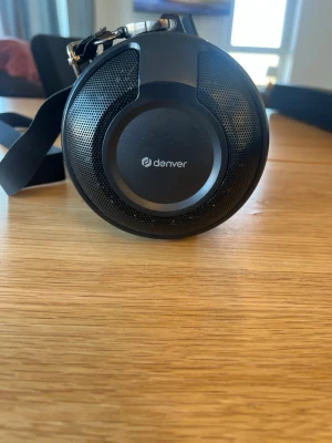 Denver bärbar högtalare svart - Bärbar högtalare från Denver i svart färg med rund form och robust design. Högtalaren har ett nätmaterial runt om och en praktisk axelrem för enkel transport. Perfekt för att ta med sig musiken överallt.