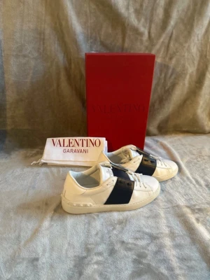 Valentino Garavani vita sneakers - Stilrena vita sneakers från Valentino Garavani med bred blå läderdetalj över sidan😁Skorna är 100%äkta då vi checkar varje produkt noggrant så dem inte är fejk 😁✅ allt ingår på köpet✅priset är inte hugget i sten och kan diskuteras 😁✅SKICK:nya 