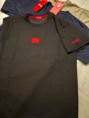 Svart t-shirt från Hugo med röd logga - Svart t-shirt från Hugo med röd logotyp på bröstet och ärmen. Klassisk passform och rund halsringning. Perfekt för en stilren och modern look.