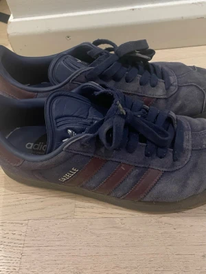 Adidas Gazelle mörkblå sneakers - Säljer ett par Adidas Gazelle sneakers i mörkblå mocka med klassiska tre ränder i vinrött på sidorna. Skorna har lågt skaft, snörning och svart sula. Perfekta för dig som gillar retrostil och vill ha något som sticker ut.