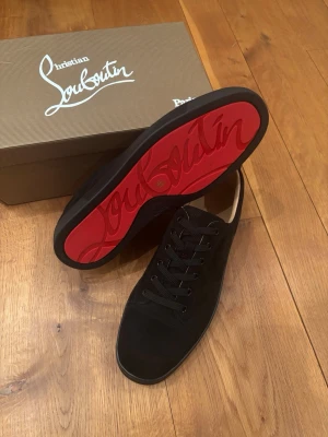 Svarta sneakers från Christian Louboutin - Stilrena svarta sneakers från Christian Louboutin med ikonisk röd sula och diskret logga. Skorna har lågt skaft, klassisk snörning och är tillverkade i mocka. Perfekta för dig som vill sticka ut med en lyxig touch.