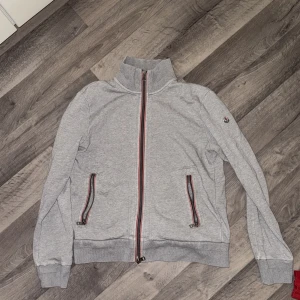 Grå kofta från Moncler - Säljer en äkta grå kofta från Moncler med ståkrage och hel dragkedja framtill. Koftan har två fickor med randiga dragkedjor och broderad Moncler-logga på ärmen. Ribbad mudd vid ärmslut och nederkant. Det än liten skavanka på vänster baksida, som är professionellt sydd av en skräddare, botten av dragkedjan där har kedjan gått av men den går fortfarande att stänga men de kan vara lite frustrerande men om man skulle vilja hade man kunnat gå till en skräddare kostar 50-150 att fixa. Finns privat🌟😇