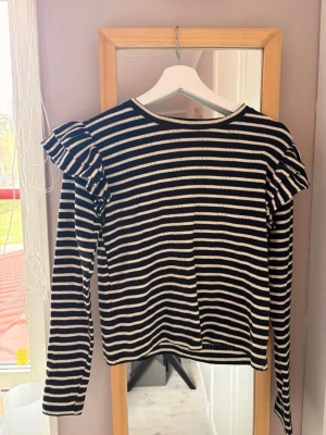 Randig långärmad topp med volang - Svart och vit randig långärmad topp med volangdetaljer på axlarna. Toppen har rund halsringning och är i en mjuk, ribbad kvalitet. Perfekt för en trendig och avslappnad look.