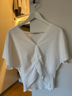 Vit omlottopp med knyt fram - Vit topp med v-ringning och omlottkänsla, knyt fram och korta ärmar. Tunt, lätt strukturerat tyg som ger en luftig känsla. Perfekt till jeans eller kjol för en fräsch look.