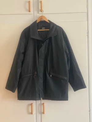 Svart parkajacka från BOSS - Stilren svart parkajacka från BOSS med hög krage och dragkedja framtill. Jackan har två snedställda fickor med dragkedja och justerbara spännen i midjan på baksidan. Materialet är slätt och jackan har en rak, något längre passform.