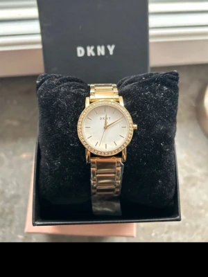 Guldfärgad DKNY klocka med kristaller - Snygg guldfärgad klocka från DKNY med klassisk rund urtavla och eleganta kristaller runt ramen. Den är i guld och har en stilren länkdesign och klockan har minimalistiska visare och index. Perfekt accessoar för dig som vill ha något stilrent och lyxigt. Den är helt oanvänd och kommer i sin original förpackning 