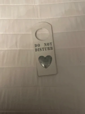 Dörrskylt Do Not Disturb med hjärta - Vit dörrskylt i rektangulär form med texten 'DO NOT DISTURB' i grått och ett grått hjärta nedtill. Skylten har ett runt hål upptill för att hängas på dörrhandtaget. Perfekt för att markera när du vill vara ifred.