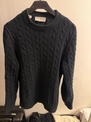 Svart kabelstickad tröja Selected - Kabelstickad svart tröja från Selected med rund halsringning och ribbade muddar. Klassisk och stilren design som passar perfekt till jeans eller chinos. Materialet känns mjukt och bekvämt, och tröjan har en snygg passform med långa ärmar.