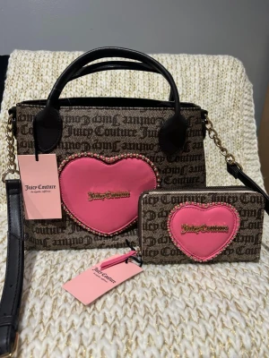 Juicy Couture brun handväska med hjärta - Juicy Couture handväska i brunt tyg med logomönster och stort rosa hjärta framtill, dekorerat med pärlor och guldfärgad logga. Svarta handtag, axelrem i svart och guld, samt matchande plånbok med samma design. Perfekt för dig som gillar statement-accessoarer.