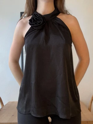 Svart halterneck topp med rosett - Snygg svart halterneck topp med en stor rosett vid halsen. Toppen är ärmlös och har en lös passform som ger en stilren och trendig look. Perfekt att matcha med jeans eller kjol för en cool outfit. Neo noir