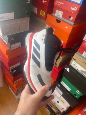 ADIDAS BOUNCE LEGENDS  vita och svarta sneakers - Snygga Adidas sneakers i vitt med svarta detaljer och röda inslag. Skorna har en sportig design med klassiska tre ränder på sidan, svart snörning och hög komfort. Ovandelen är i mesh och syntetmaterial för bra andningsförmåga och stilren look.