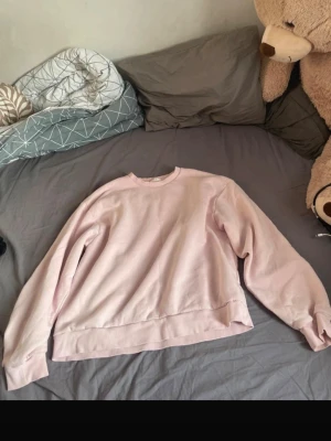 Ljusrosa sweatshirt utan huva - En basic ljusrosa sweatshirt med rund hals och långa ärmar. Tröjan har en avslappnad passform och ribbade muddar vid ärmslut och nederkant. Perfekt för en chill och stilren look.