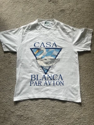 Vit Casa Blanca t-shirt med svan - Vit t-shirt från Casa Blanca med stor tryckt svan och blått grafiskt mönster på bröstet. Texten 'Casa Blanca Par Avion' i blått framtill. Klassisk passform och rund hals. Perfekt för dig som gillar statement-plagg.