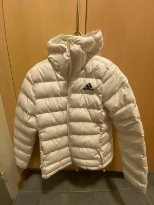 Vit pufferjacka från Adidas - Snygg vit pufferjacka från Adidas med huva och dragkedja framtill. Jackan har det klassiska Adidas-loggan på bröstet och breda, vadderade paneler som ger en riktigt fluffig look.