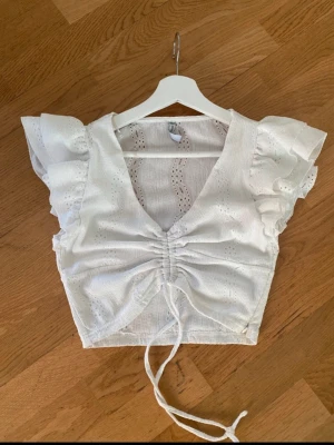 Vit blus med volangärmar från FB Sister - Supersöt vit blus från FB Sister med volangärmar, v-ringning och snörning framtill. Blusen har broderade detaljer och är croppad, perfekt till ett par höga jeans eller kjol. Lätt och luftig, passar perfekt till varma dagar.