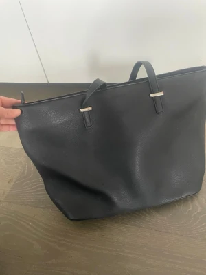 Svart toteväska i skinnimitation - Stilren svart toteväska i skinnimitation med två handtag och diskreta metalldetaljer.Perfekt att bära över axeln eller i handen. Perfekt till skolan då den rymmer mycket. Lite sliten vid handtagen men inget som syns mycket