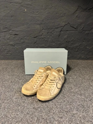Philippe Model vita sneakers  - Snygga vita sneakers från Philippe Model i läder med beige detaljer och svart hälparti. Klassisk låg modell med rund tå, vita skosnören och den ikoniska sköldloggan på sidan. Perfekta för en avslappnad och trendig stil.