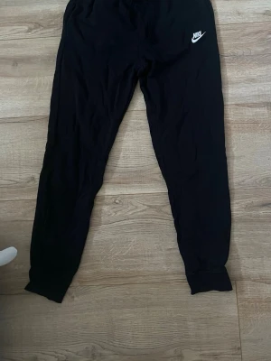 Svarta mjukisbyxor från Nike - Säljer ett par svarta mjukisbyxor från Nike med klassisk vit logga på vänster sida. Byxorna har resår i midjan och muddar vid bensluten för en skön passform. Perfekta för chill dagar eller träning. Materialet känns mjukt och bekvämt.