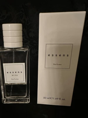 Essens W196 Parfym 50 ml - Minimalistisk parfym från Essens, modell W196. Flaskan är rektangulär i genomskinligt glas med vit etikett och vit kork. Förpackningen är stilren och vit med svart text. Innehåller 50 ml.