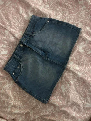 Blå jeanskjol med fickor - Säljer en klassisk blå jeanskjol i kort modell med fem fickor och bälteshällor. Kjolen har knapp och dragkedja framtill och är gjord i ett slitstarkt denimtyg. Perfekt att styla med en enkel t-shirt eller hoodie.