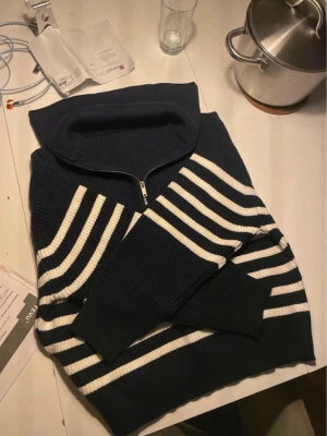 Marinblå stickad tröja med dragkedja - Säljer en marinblå stickad tröja med vita horisontella ränder och halv dragkedja vid kragen. Tröjan har en bred krage och ribbade muddar vid ärmslut och nederkant. Perfekt för dig som gillar klassisk och avslappnad stil.