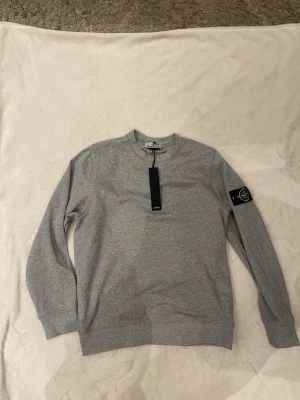 Grå sweatshirt från Stone Island - Snygg grå sweatshirt från Stone Island med klassisk rund hals och ikonisk svart patch med logga på vänster ärm. Tröjan har ribbade muddar vid ärmslut och nederkant. Storlek XXL passar även XL och L om man gillar tröjorna lösa. Perfekt för en clean och stilren look. NFC finns. Skickas samma dag!! Kan köra lite billigare vid snabb affär. Helt ny aldrig använd.