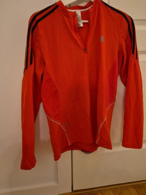 Röd långärmad träningströja Adidas - Snygg röd långärmad träningströja från Adidas med halv dragkedja framtill och svarta ränder på axlarna. Tillverkad i ett lätt och ventilerande funktionsmaterial, perfekt för löpning eller gym. Reflekterande detaljer och klassisk Adidas-logga på bröstet.