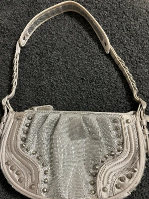 Silvrig axelremsväska med nitar - Snygg handväska i metallic-silver med glittrande material och dekorativa nitar framtill. Väskan har en flätad axelrem och dragkedja upptill.
