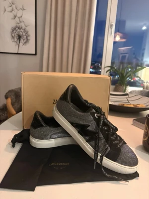 Svarta sneakers från Zadig & Voltaire - Snygga svarta sneakers från Zadig & Voltaire med glittrig textil, svarta läderdetaljer och vit sula. Skorna har en cool blixtdetalj på sidan och svarta skosnören med text. Perfekta för dig som vill sticka ut med din stil. Köpte de för 2500kr.