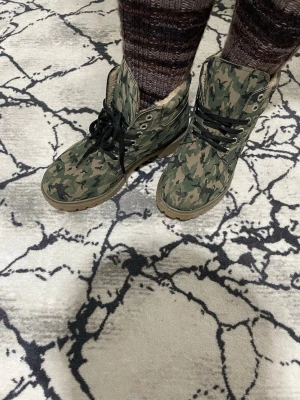 Camo mönstrade kängor med snörning - Säljer ett par kängor med grönt camo-mönster och svarta snören. De har grov sula i gummi och högt skaft med metallöljetter. Insidan verkar vara fodrad för extra komfort. Perfekta för dig som vill sticka ut med en tuff stil.