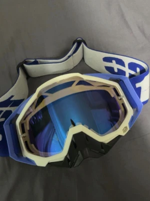 Blå motocrossglasögon / skidglasögon  - Snygga motocrossglasögon/skidglasögon vet ej vart de är ifrån. Med blått och vitt band, blått yttre plast och vit ram. De har en robust design med svart nässkydd och bred elastisk rem för bra passform. Passar min skidhjälm i S