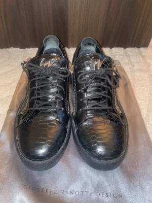 Svarta sneakers Giuseppe Zanotti Box/Dustbag - Snygga svarta sneakers från Giuseppe Zanotti med blank yta och ormmönstrad struktur. Skorna har guldfärgade dragkedjor på sidorna och klassiska svarta skosnören. Ikonisk logga på plösen och robust sula för en lyxig streetwear-look.