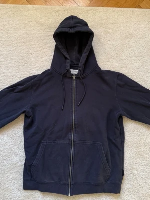 Mörkblå hoodie med dragkedja Pier One - Klassisk mörkblå hoodie från Pier One med dragkedja framtill, två stora fickor och justerbar huva med snören. Tillverkad i mjukt material som känns skönt mot huden. Perfekt för en avslappnad och stilren look.