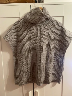 Grå stickad poloväst - Säljer en grå stickad väst med hög polokrage och korta ärmar. Västen har ett grovt ribbstickat mönster och är perfekt att slänga över en skjorta eller långärmad tröja. Materialet känns mjukt och mysigt, men exakt material är okänt.