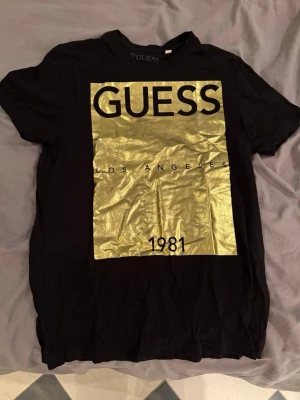 Svart t-shirt Guess med guldtryck - Snygg svart t-shirt från Guess med stort guldigt tryck på framsidan där det står 'Guess Los Angeles 1981'. Klassisk passform och korta ärmar, perfekt för en trendig och avslappnad stil. Materialet känns mjukt och bekvämt mot huden.