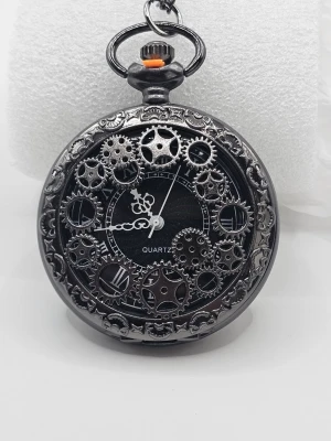 Svart steampunk fickur med kugghjul "klocka" - Unikt svart fickur i steampunk-stil med detaljerade kugghjul på urtavlan och dekorativ ram. Klockan har vita visare och romerska siffror, samt en kedja för att fästa i fickan. Perfekt accessoar för dig som gillar retro och industriell design.