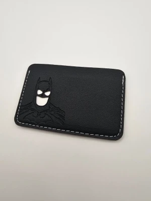 Svart Batman kortfodral i läder - Säljer ett svart kortfodral i läder med Batman-motiv präglat på framsidan. Fodralet har vita sömmar runt kanterna och är perfekt för att förvara kort som körkort eller bankkort. Stilren och cool design för dig som gillar superhjältar.