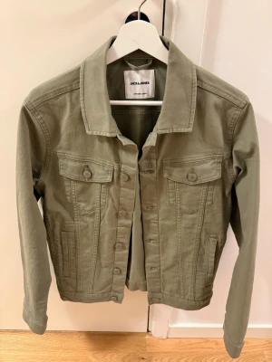 Olivgrön jeansjacka från Jack & Jones - Stilren olivgrön jeansjacka från Jack & Jones med klassisk krage, två bröstfickor med lock och knappar samt raka ärmar. Jackan har en snygg, enkel design och är tillverkad i jeansmaterial.