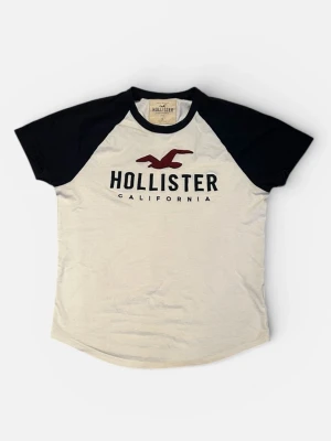 Vit och svart Hollister t-shirt - Snygg t-shirt från Hollister med svart-vita kontraster och klassisk logga på bröstet. T-shirten har svarta korta ärmar och vit kropp, samt rund halsringning. Tillverkad i mjuk bomull för skön känsla och perfekt passform.