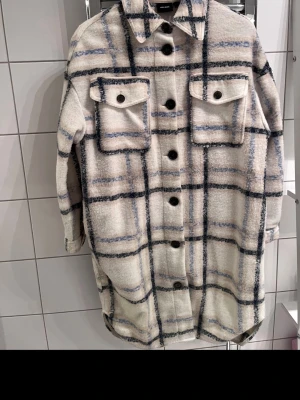 Rutig overshirt från Vero Moda - Overshirt i beige och blått rutmönster från Vero Moda. Modellen har stora bröstfickor med lock, svarta knappar framtill och en klassisk krage. Materialet är mjukt och borstat, perfekt för lager på lager. Lång och avslappnad passform.