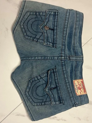 Blå jeansshorts från True Religion - Säljer ett par blå jeansshorts från True Religion med ikoniska kontrastsömmar och stora fickor bak. Shortsen har flera metallknappar framtill och klassisk patch på baksidan. Perfekta för sommaren och har en snygg, casual look.