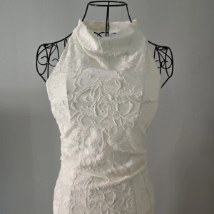 Vit lång spetsklänning med hög hals - Elegant vit långklänning med hög hals och ärmlös design. Klänningen har vackra broderade blommor i spets och en figurnära passform som ger en stilren siluett. Helt oanvänd, Prislappen finns kvar 