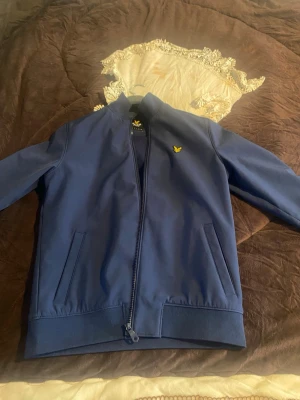 Blå  Lyle & Scott jacka - Snygg marinblå bomberjacka från Lyle & Scott med gul logga på bröstet. Jackan har dragkedja framtill, ribbade muddar och två sidofickor. Perfekt för dig som vill ha en stilren och enkel jacka till vardags.
