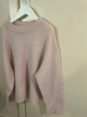 Ljusrosa stickad tröja Gina Tricot -  ljusrosa stickad tröja från Gina Tricot med bred ribbad kant vid hals, ärmslut och nederkant. Tröjan har en avslappnad passform och är perfekt för att hålla värmen under kyliga dagar, och för en tröja till vardagen.