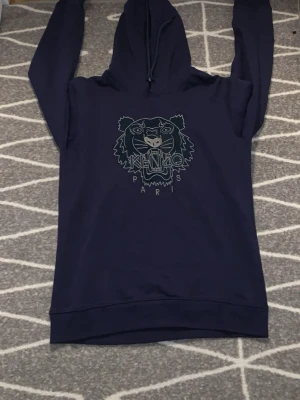 Mörkblå Kenzo hoodie med tigertryck - Snygg mörkblå hoodie från Kenzo med det ikoniska tigerhuvudet och Kenzo Paris-tryck på bröstet. Klassisk huva med dragsko och långa ärmar. Tillverkad i mjukt material som känns skönt mot huden. Perfekt för dig som vill ha en streetwear vibe.