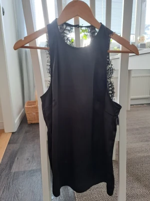 Svart ärmlös topp med spetsdetaljer - Snygg svart ärmlös topp med spetsdetaljer längs axlar och rygg. Toppen har en rund halsringning och stängs med en liten knapp i nacken. 