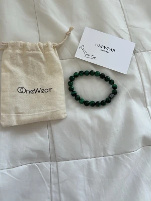 Grönt pärlarmband från OneWear - Snyggt armband från OneWear med mörkgröna pärlor och en metallisk detalj. Armbandet är elastiskt och består av runda pärlor som ger en stilren och modern look. Perfekt accessoar för att lyfta din outfit.