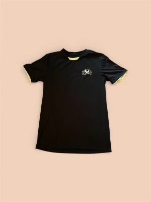 Svart tränings-tshirt från Soc - Svart tränings-tshirt från Soc med gul logga och grafiskt tryck på bröstet. T-shirten har gula detaljer vid ärmslut och halsringning. Tillverkad i ett lätt och snabbtorkande material som passar perfekt för gym eller löpning.