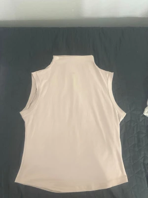 Beige ärmlös polotopp - Säljer en stilren beige polotopp utan ärmar. Toppen har en hög krage och är tillverkad i ett mjukt material som känns skönt mot huden. Perfekt att matcha med jeans eller kjol för en clean look.