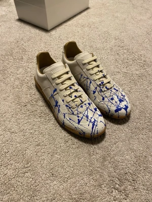 Maison Margiela Blue Paint Splatter Gats - Unika sneakers från Maison Margiela i vitt läder med beige detaljer och blå färgstänk över hela ovandelen. Klassisk låg modell med gummisula och tonade skosnören. Perfekt för dig som vill sticka ut med en kreativ och modern look. True to size